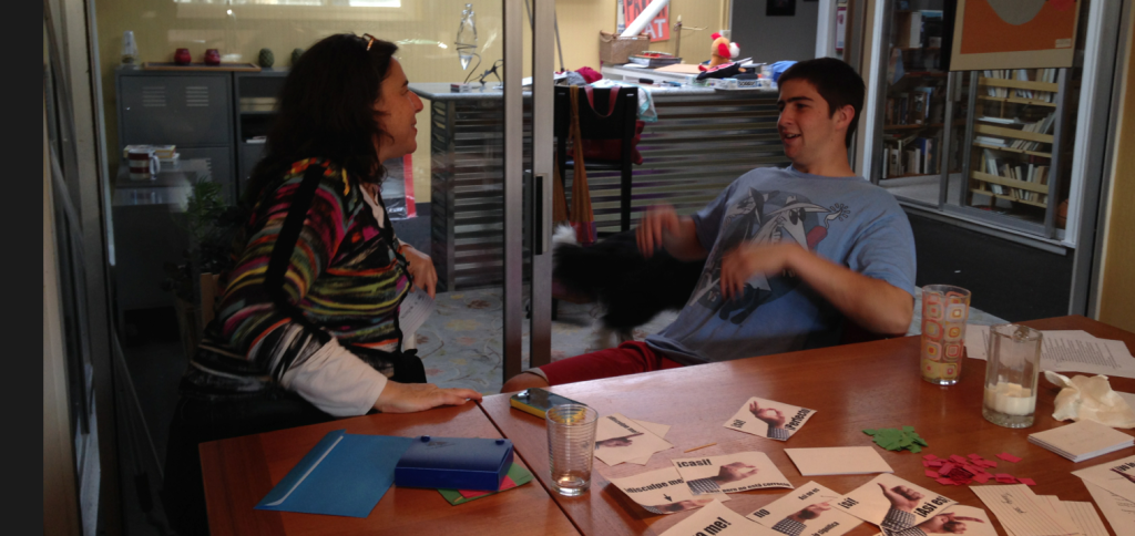 Kid gestures, smiling, while mom guesses. In the foreground, cards with pictures of hands are on the table. 